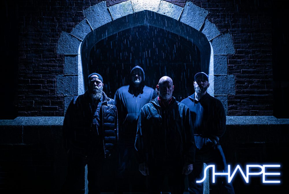 The band SHAPE: four members standing in the rain under a lit stone archway at night.